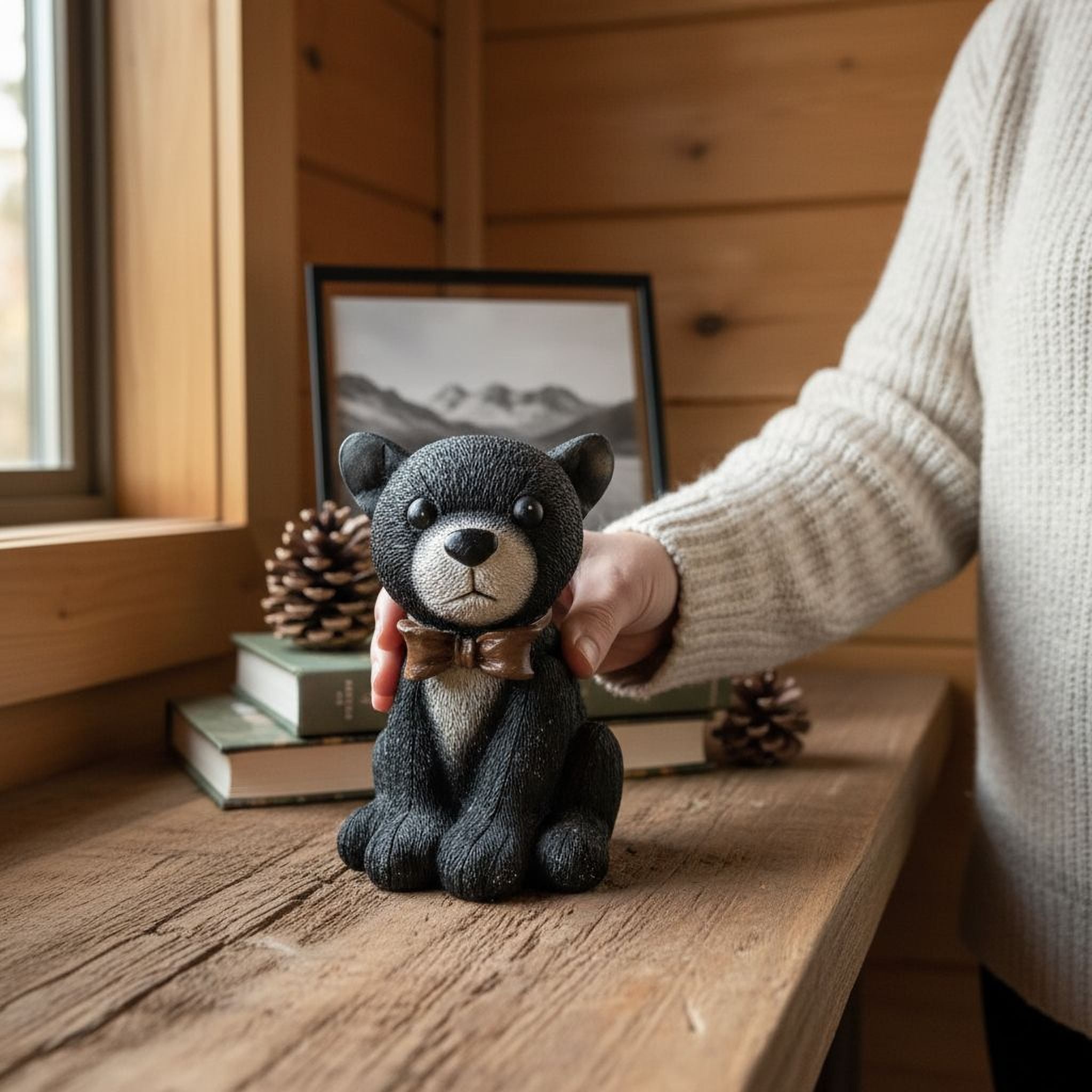 A person is holding a small black bear figurine with a bow tie on a wooden surface, with a framed picture and pine cones in the background.