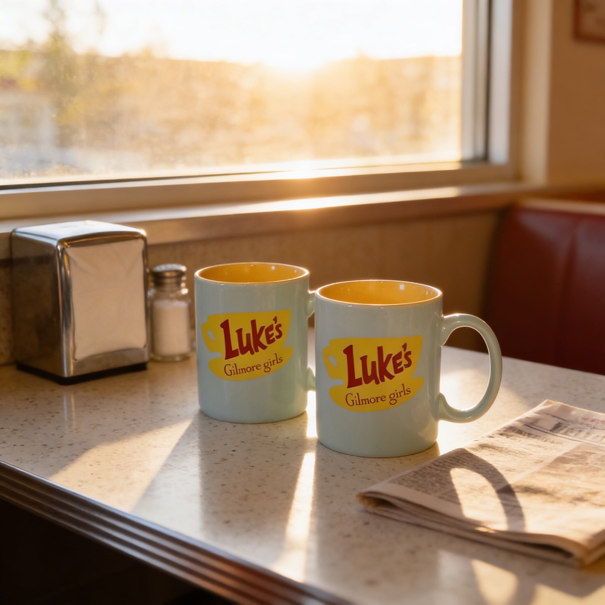Two light blue mugs with yellow text and a logo, placed on a table near a window and a newspaper.