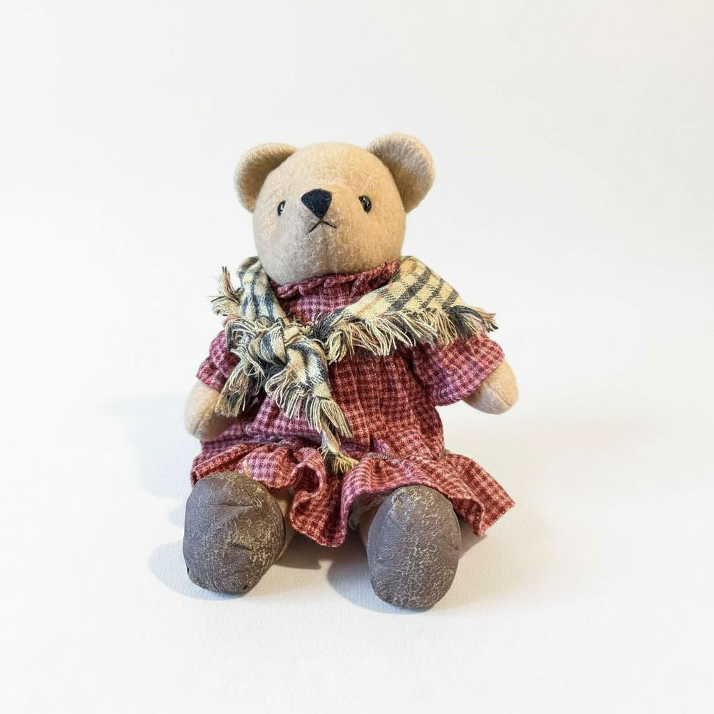 A brown teddy bear wearing a red and white checkered dress and a plaid scarf, sitting on a plain white background.