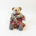 A brown teddy bear wearing a red and white checkered dress and a plaid scarf, sitting on a plain white background.