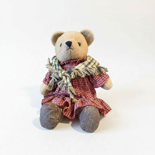 A brown teddy bear wearing a red and white checkered dress and a plaid scarf, sitting on a plain white background.