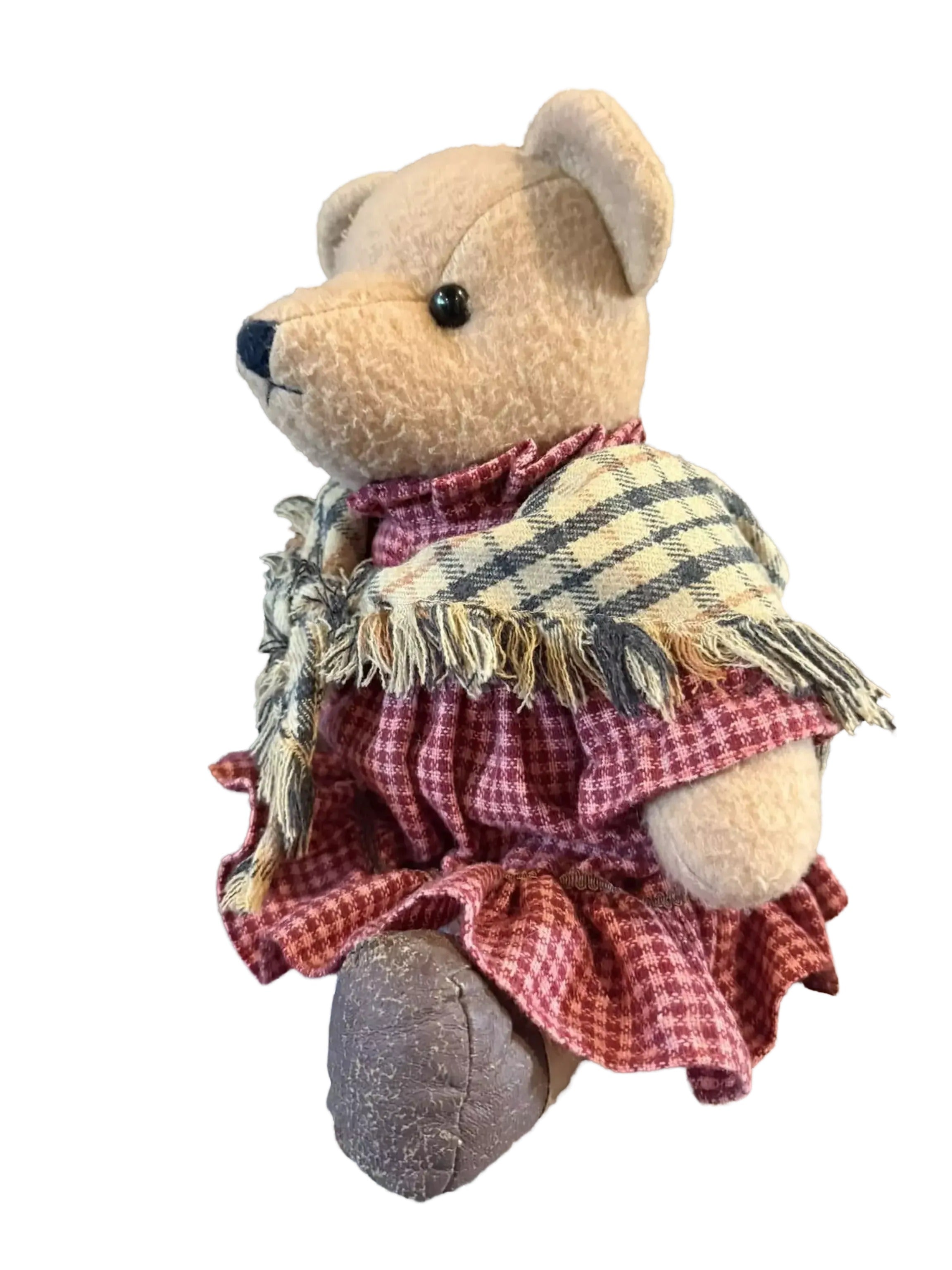A brown teddy bear wearing a red and white checkered dress sits on a gray surface, with a stone wall in the background.