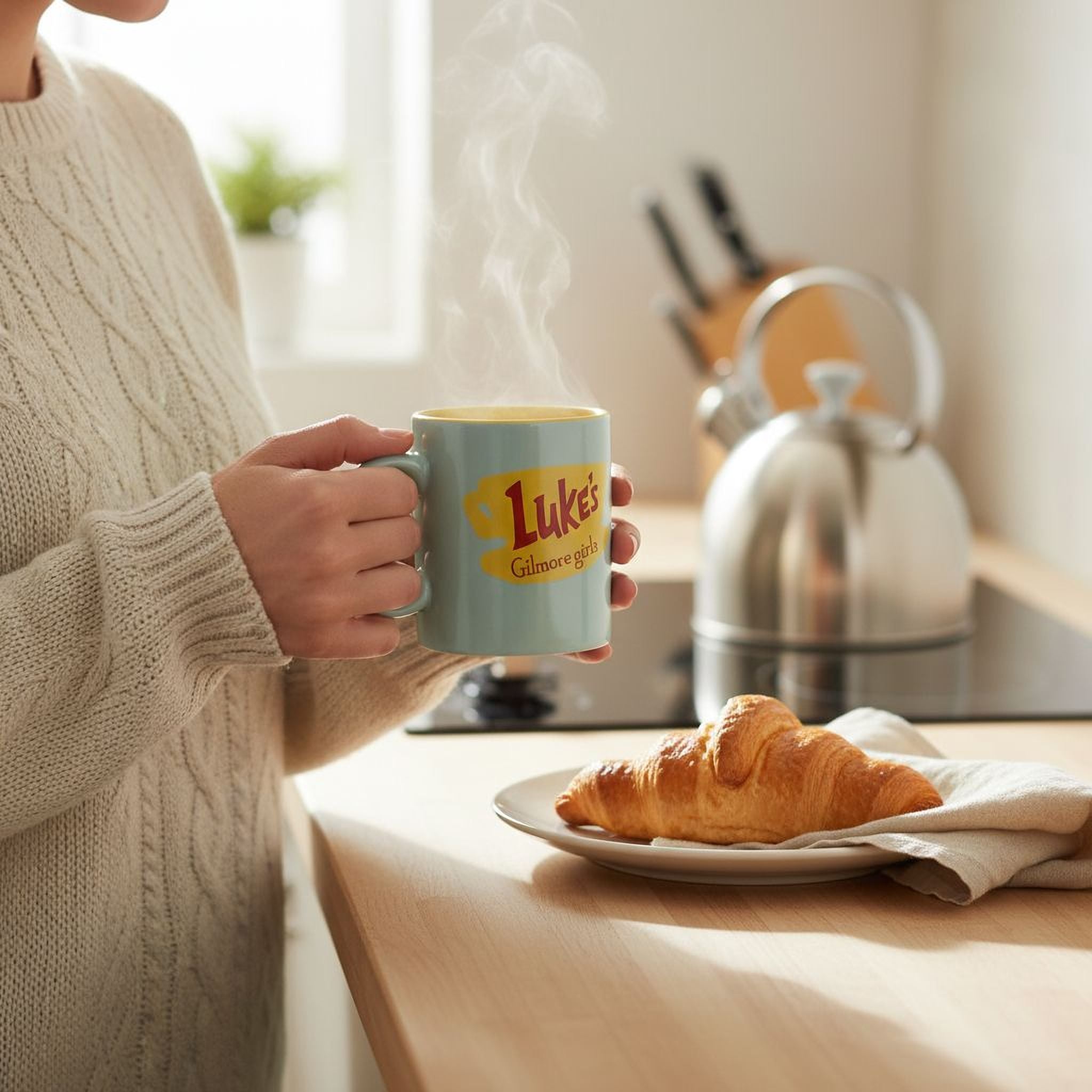 A person is holding a mug with the text " Luke's GIRL" on it, while standing in a kitchen with a plate of food and a kettle on the counter.